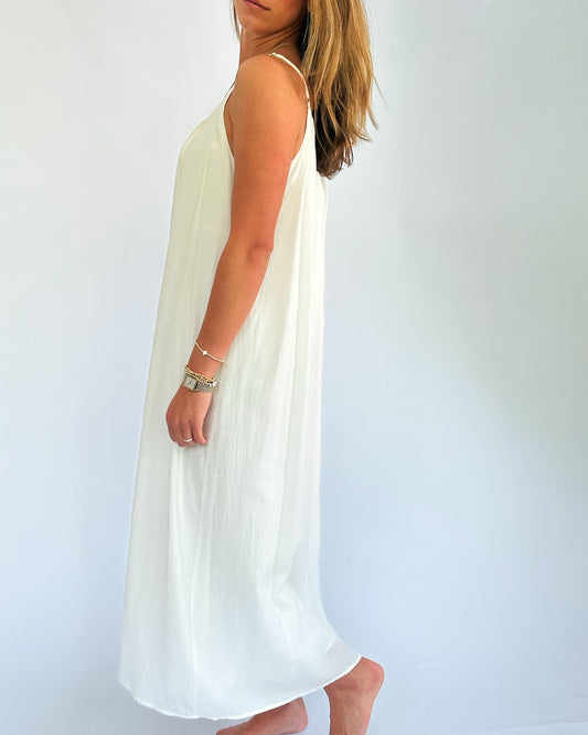Woman in a flowing white dress, standing barefoot against a plain background. She gazes sideways, exuding a calm, relaxed vibe.