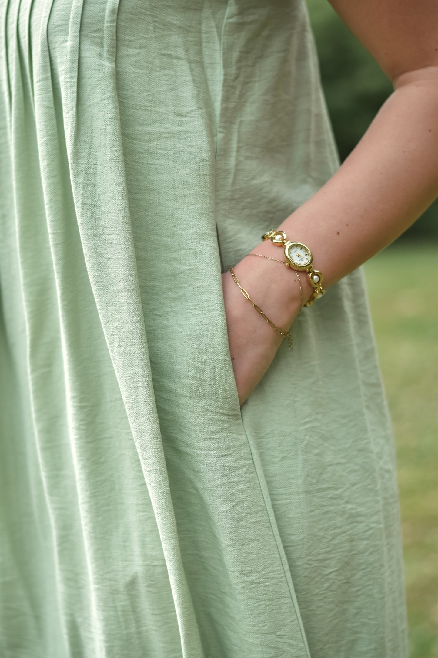 Close-up of a person wearing a light green dress with their hand in the pocket. They have a vintage-style bracelet watch. The tone is relaxed and elegant.
