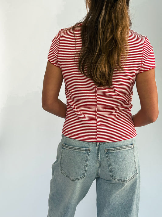 A woman with long hair stands facing away, wearing a red and white striped top and light blue jeans, conveying a casual, relaxed tone.