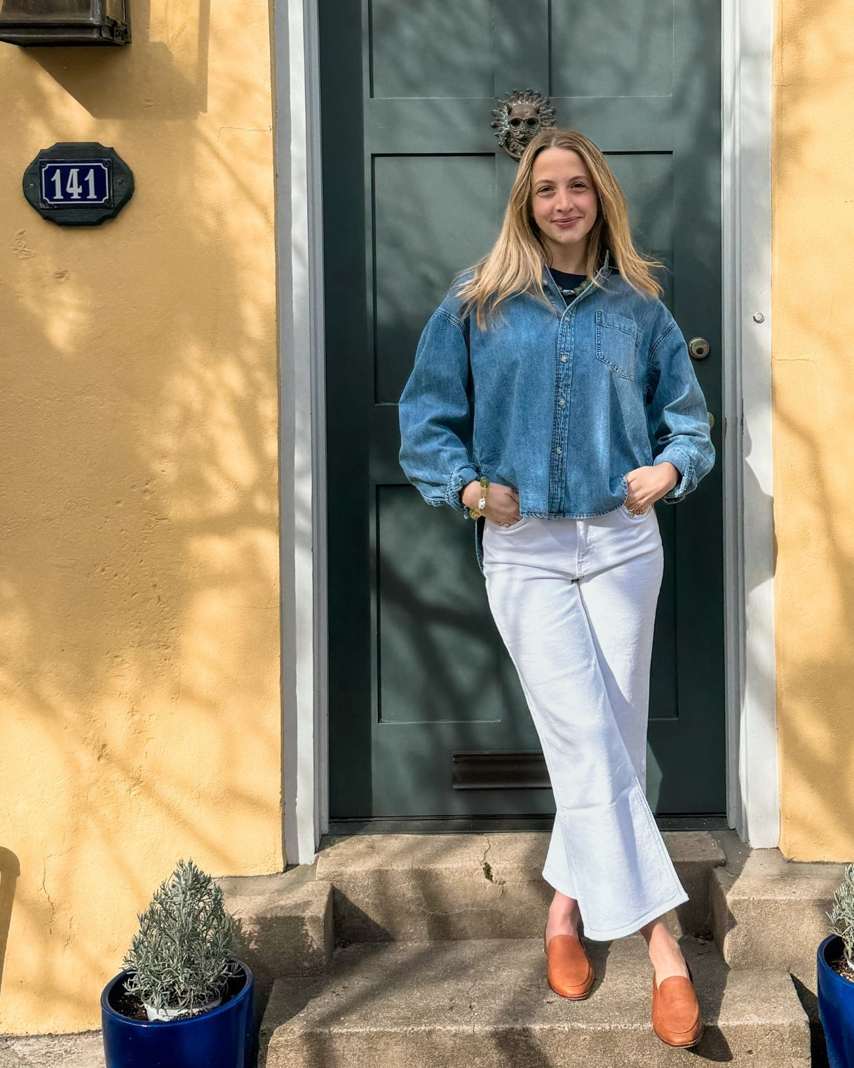 A person stands confidently in front of a green door, wearing a denim jacket and white pants. Sunlight casts soft shadows on the yellow wall.