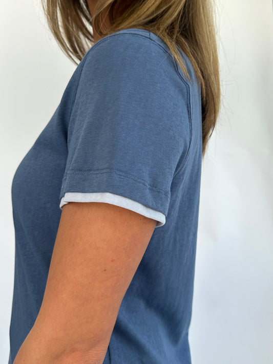 A person wearing a blue short-sleeved T-shirt is shown from the shoulders down against a white background. The outfit is casual and relaxed.