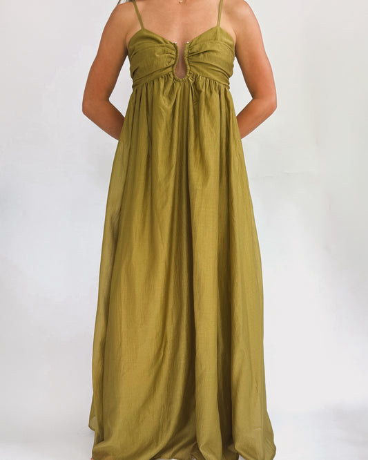 A person wearing a long, olive green dress stands in front of a plain white background. The dress has thin straps and a gathered bust, exuding elegance.