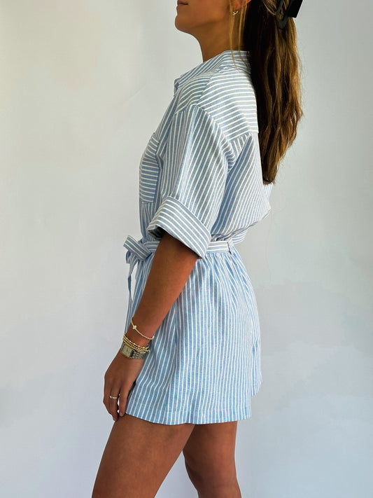 Woman in a side profile wearing a light blue and white striped romper with rolled sleeves and a waist tie. Her hair is long and features a black clip.