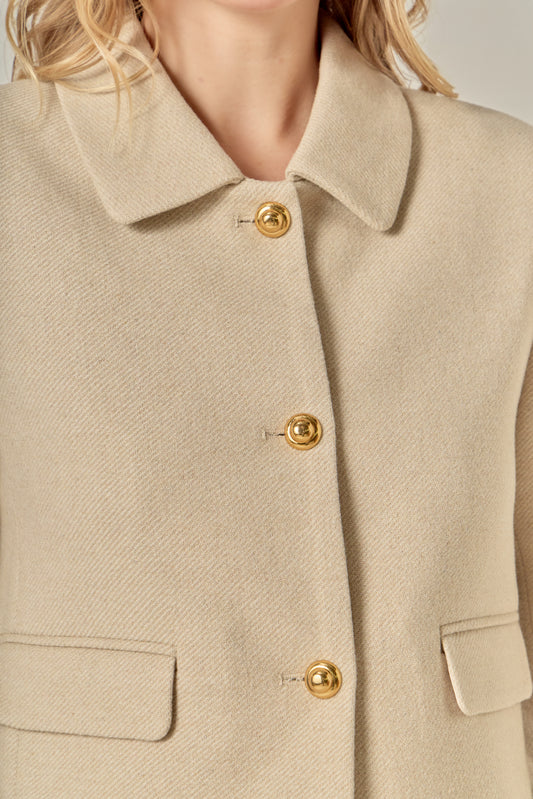 A woman with wavy blonde hair wears a beige buttoned jacket and matching shorts.