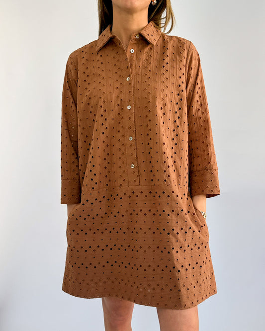 A person in a rust colored, patterned button-up dress with three-quarter sleeves, standing with hands in pockets against a plain white background.