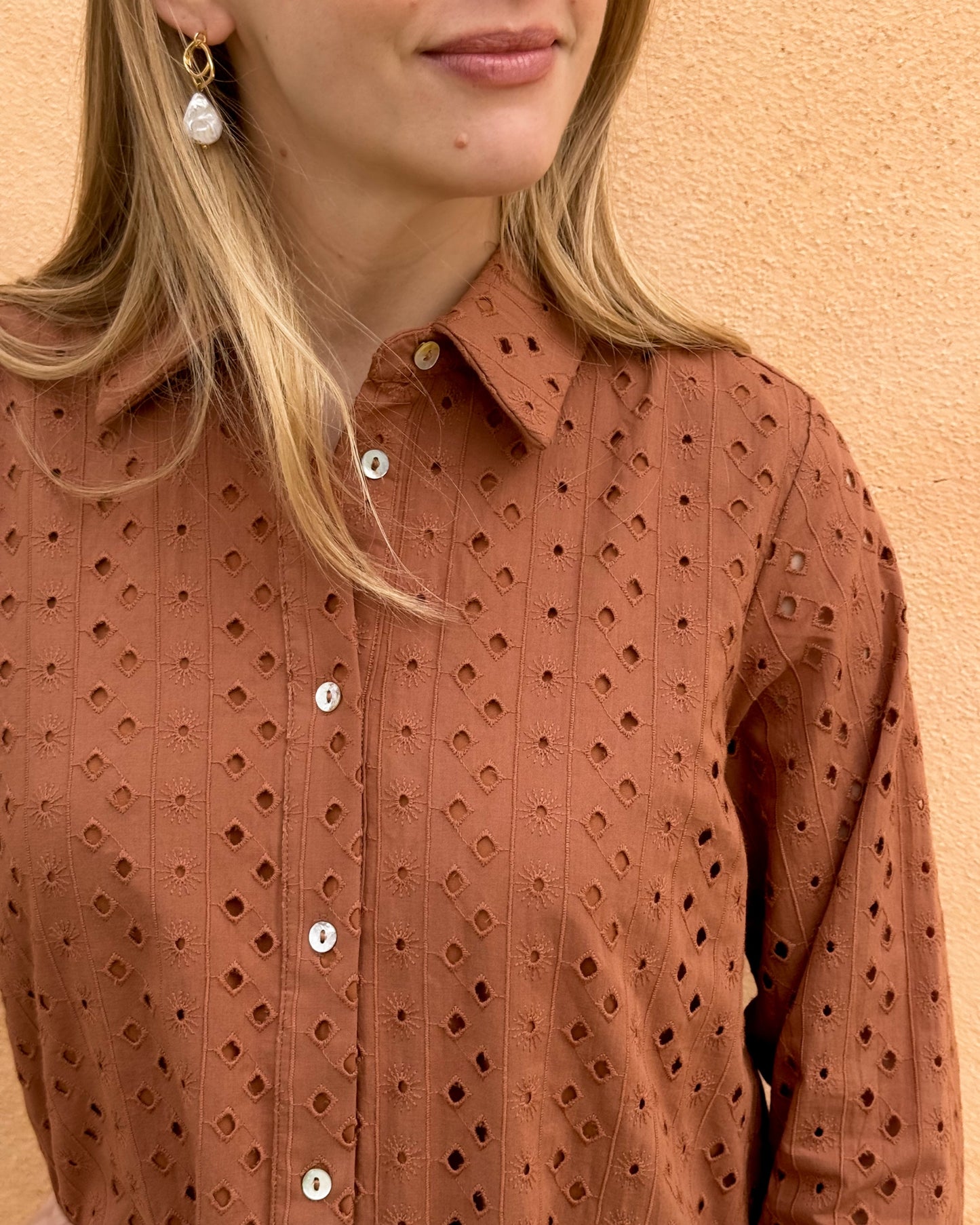 A woman stands against a textured, tan wall wearing a rust-colored, eyelet-patterned dress with white buttons.