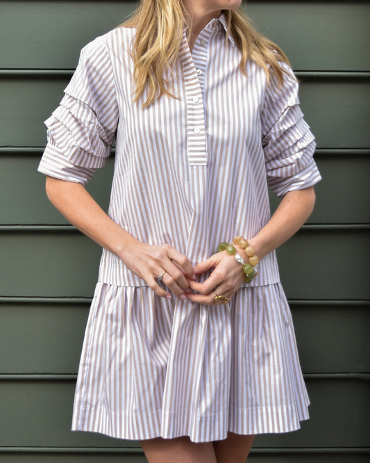 Woman in a striped shirt with puffed sleeves stands against a gray background. Her blonde hair is down, and she wears pearl earrings. The mood is casual.
