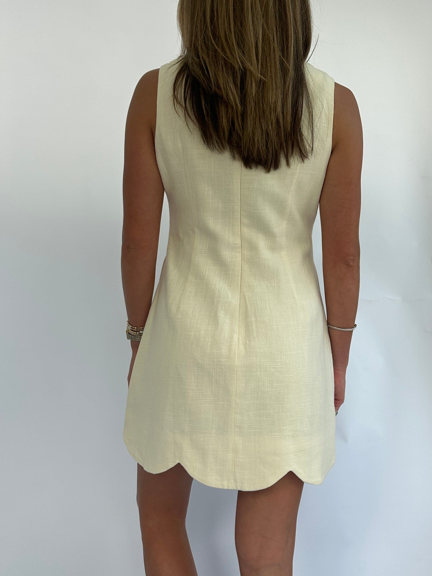 Rear view of a woman in a sleeveless, butter yellow-colored dress with a scalloped hem, standing against a plain background. Her straight hair falls mid-back.