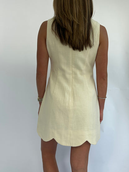 Rear view of a woman in a sleeveless, butter yellow-colored dress with a scalloped hem, standing against a plain background. Her straight hair falls mid-back.