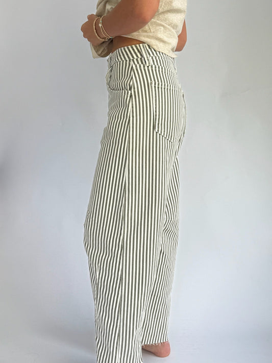 A person wearing cream and olive vertically striped barrel jeans and a beige shirt is holding the shirt hem. The background is plain light gray.