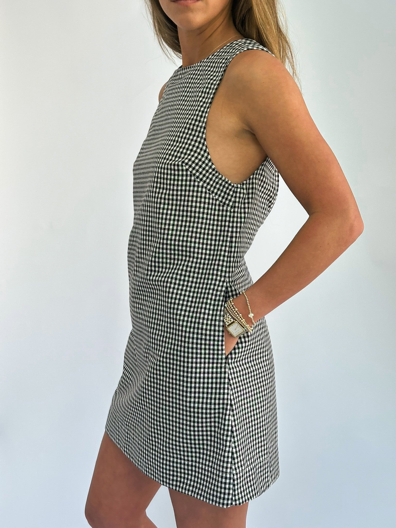 A woman stands against a plain background, wearing a sleeveless, black-and-white checkered dress. Her hand is on her hip, displaying bracelets and a watch.