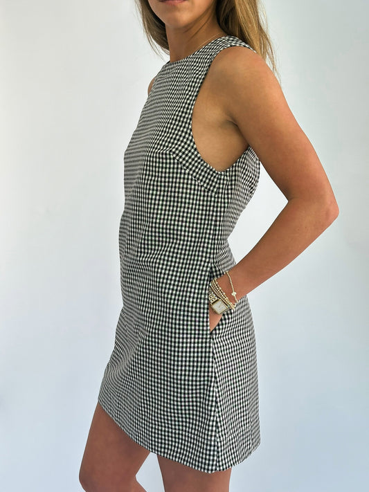 A woman stands against a plain background, wearing a sleeveless, black-and-white checkered dress. Her hand is on her hip, displaying bracelets and a watch.