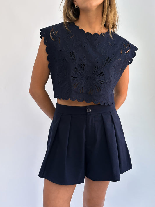 A person wears a navy blue lace-patterned sleeveless crop top with scalloped edges, paired with high-waisted shorts. The style is casual and elegant.