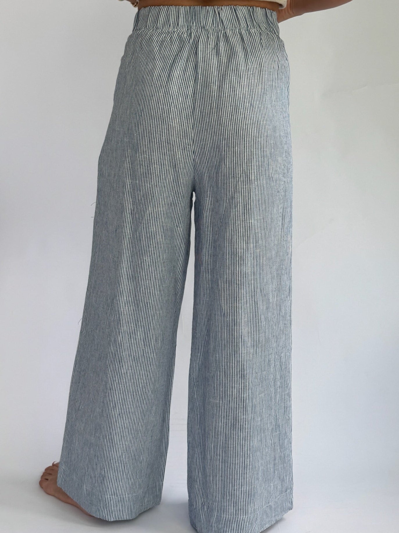 Person wearing high-waisted, blue and white striped, wide-leg linen pants with buttons. Hands on hips; casual, relaxed tone. Barefoot on plain background.