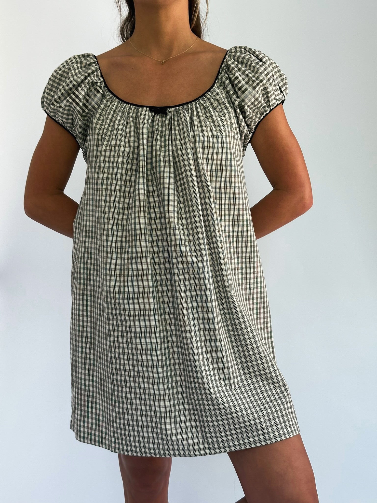 Woman in a checkered dress with puff sleeves and black trim, standing against a plain background. Her hands are behind her back, exuding a relaxed vibe.