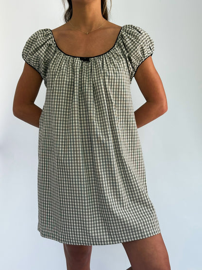 Woman in a checkered dress with puff sleeves and black trim, standing against a plain background. Her hands are behind her back, exuding a relaxed vibe.