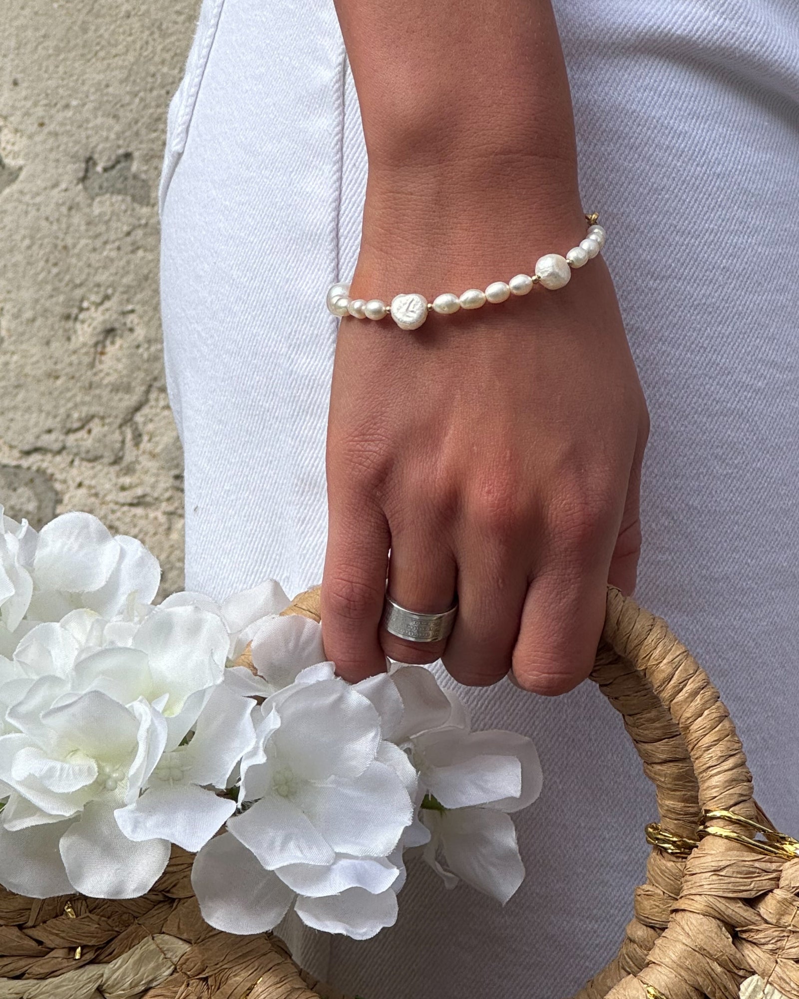 A hand with a silver ring and pearl bracelet holds a straw basket filled with white flowers. The person is wearing white pants against a stone wall. The tone is elegant and serene.