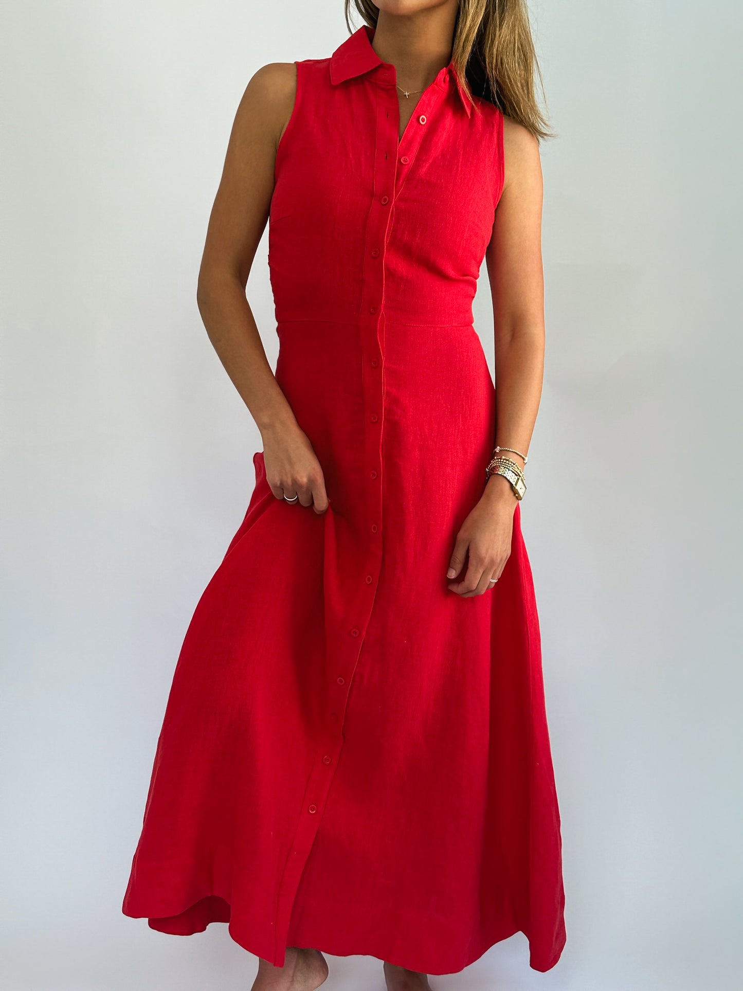 A woman in a sleeveless, button-front red dress stands with hands in her pockets. She wears bracelets and her hair is down, conveying a casual, elegant vibe.