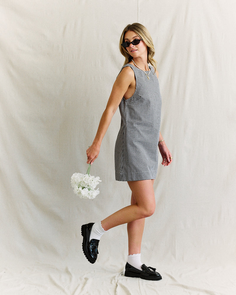 A woman in a checkered dress and black loafers poses with her right hand holding white flowers. She wears sunglasses, exuding a chic, relaxed vibe.