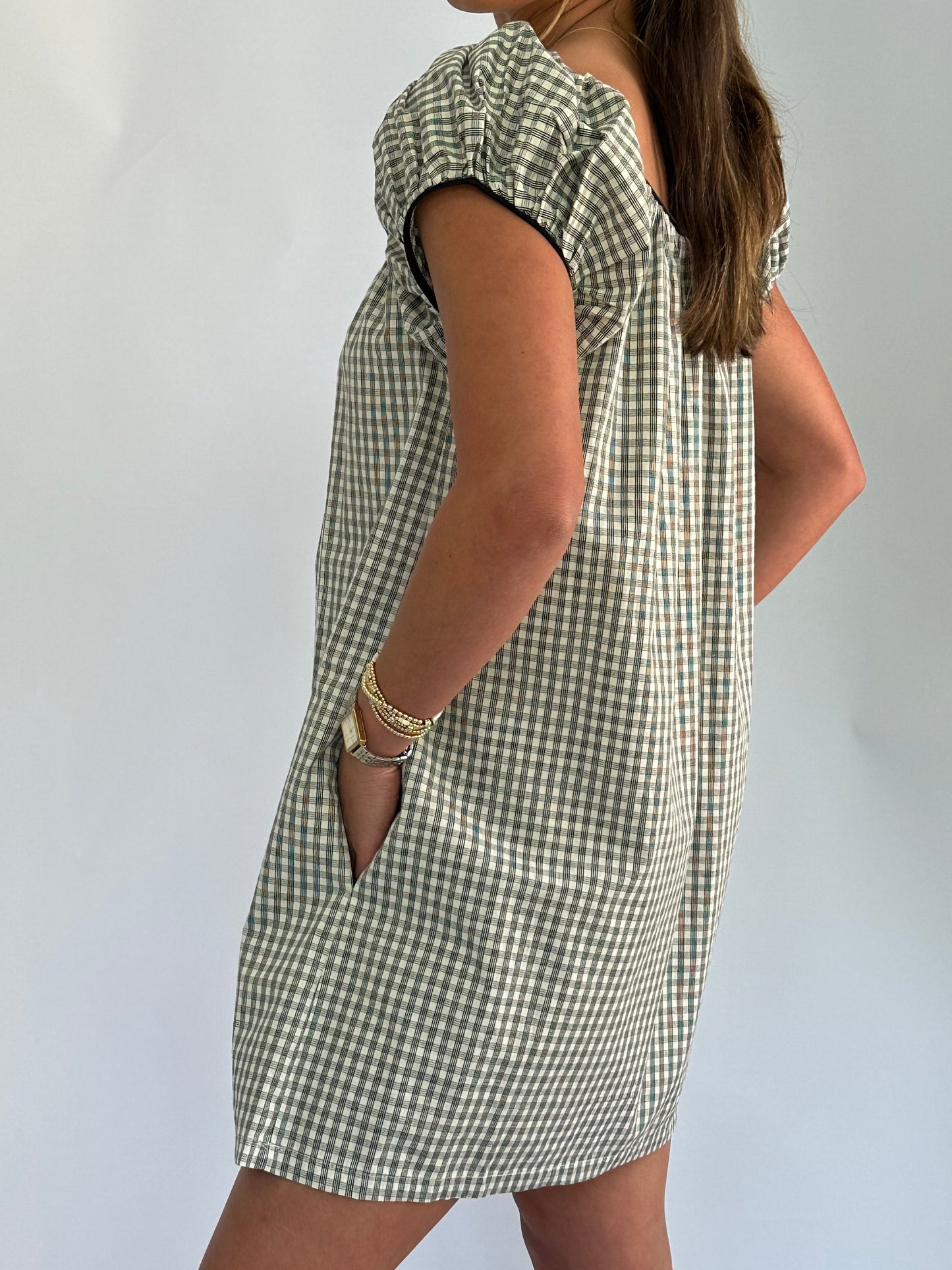 Woman stands sideways, wearing a short-sleeved, checkered dress with hands in pockets. Her long hair is loose, and she wears gold bracelets.