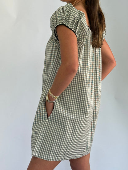 Woman stands sideways, wearing a short-sleeved, checkered dress with hands in pockets. Her long hair is loose, and she wears gold bracelets.