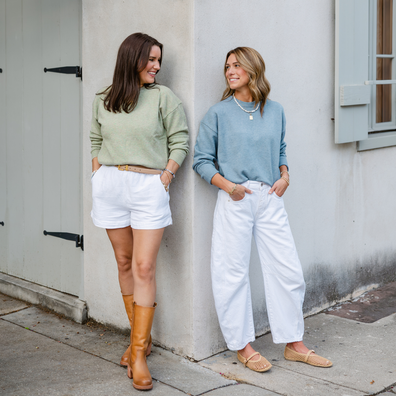 Two women stand against a corner wall, smiling at each other. One wears a green sweater, white shorts, and brown boots. The other wears a blue sweater, white pants, and woven flats, creating a casual and cheerful vibe.