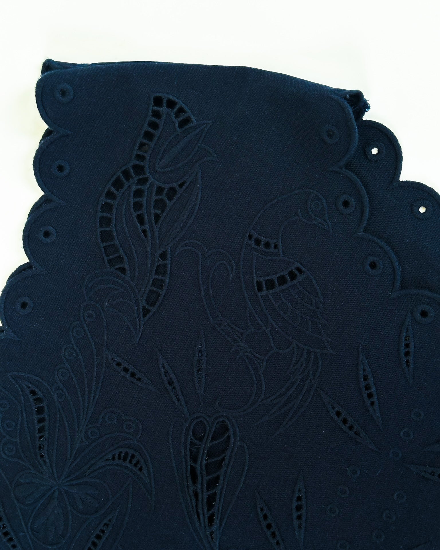 Close-up of dark fabric featuring intricate floral and bird embroidery with cut-out patterns and scalloped edges, conveying elegance and craftsmanship.