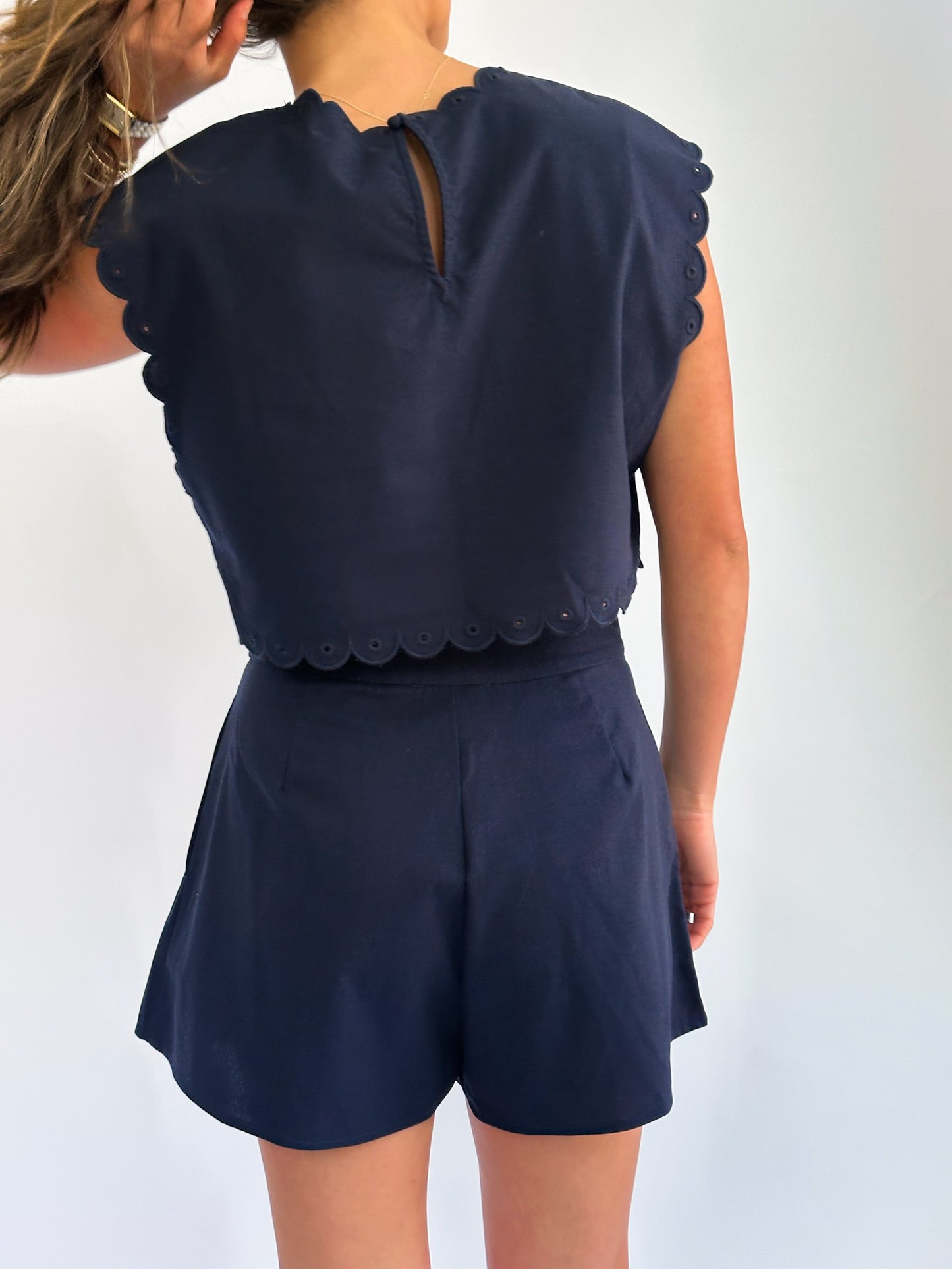 Rear view of a woman in a navy blue outfit with scalloped edges, featuring a sleeveless top and shorts. She holds her wavy hair, conveying a casual vibe.