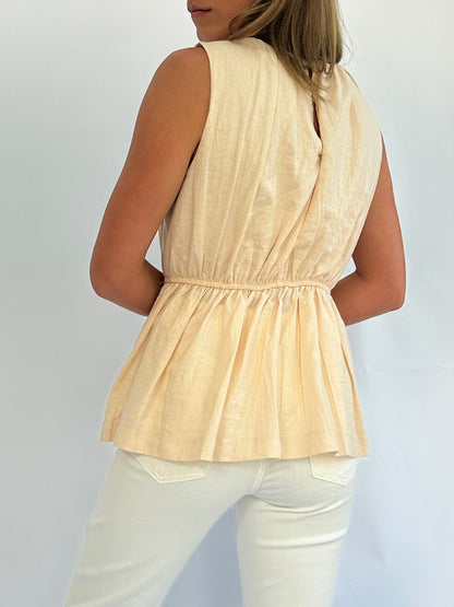 A person wearing a sleeveless, peach-colored, gathered top. The outfit features a cinched waist and is paired with light-colored pants, conveying a casual and elegant style.