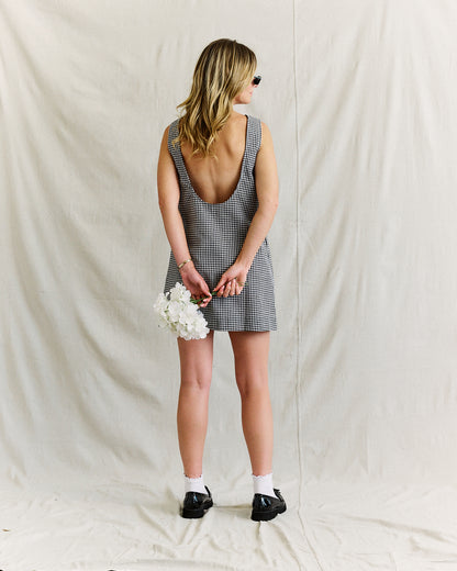 A woman stands against a cream backdrop, wearing a sleeveless, backless checkered dress. She holds white flowers, wearing sunglasses, socks, and loafers.