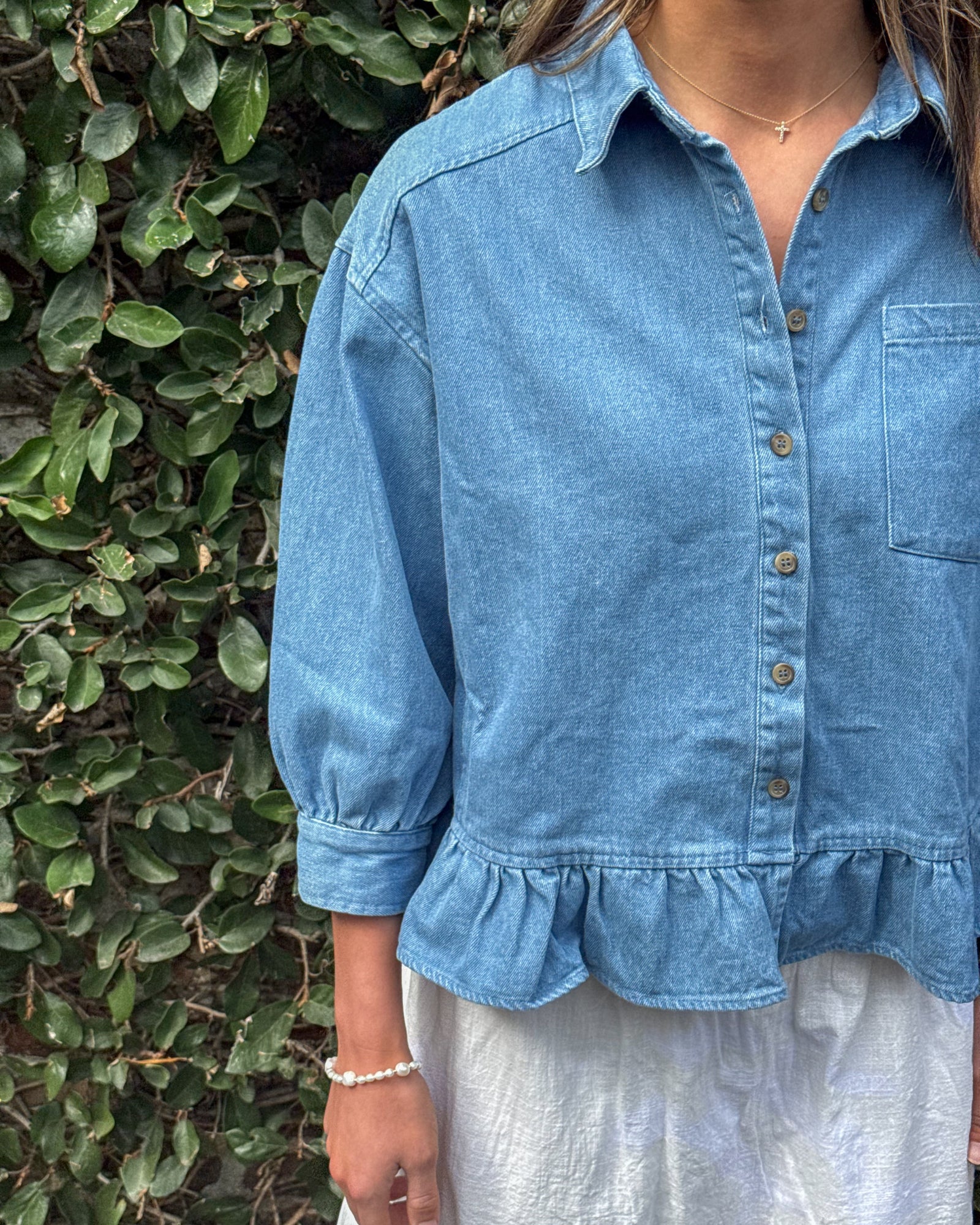 A woman in a blue denim blouse with ruffled hem stands against a leafy background. She wears a delicate necklace and bracelet, conveying a casual, natural vibe.