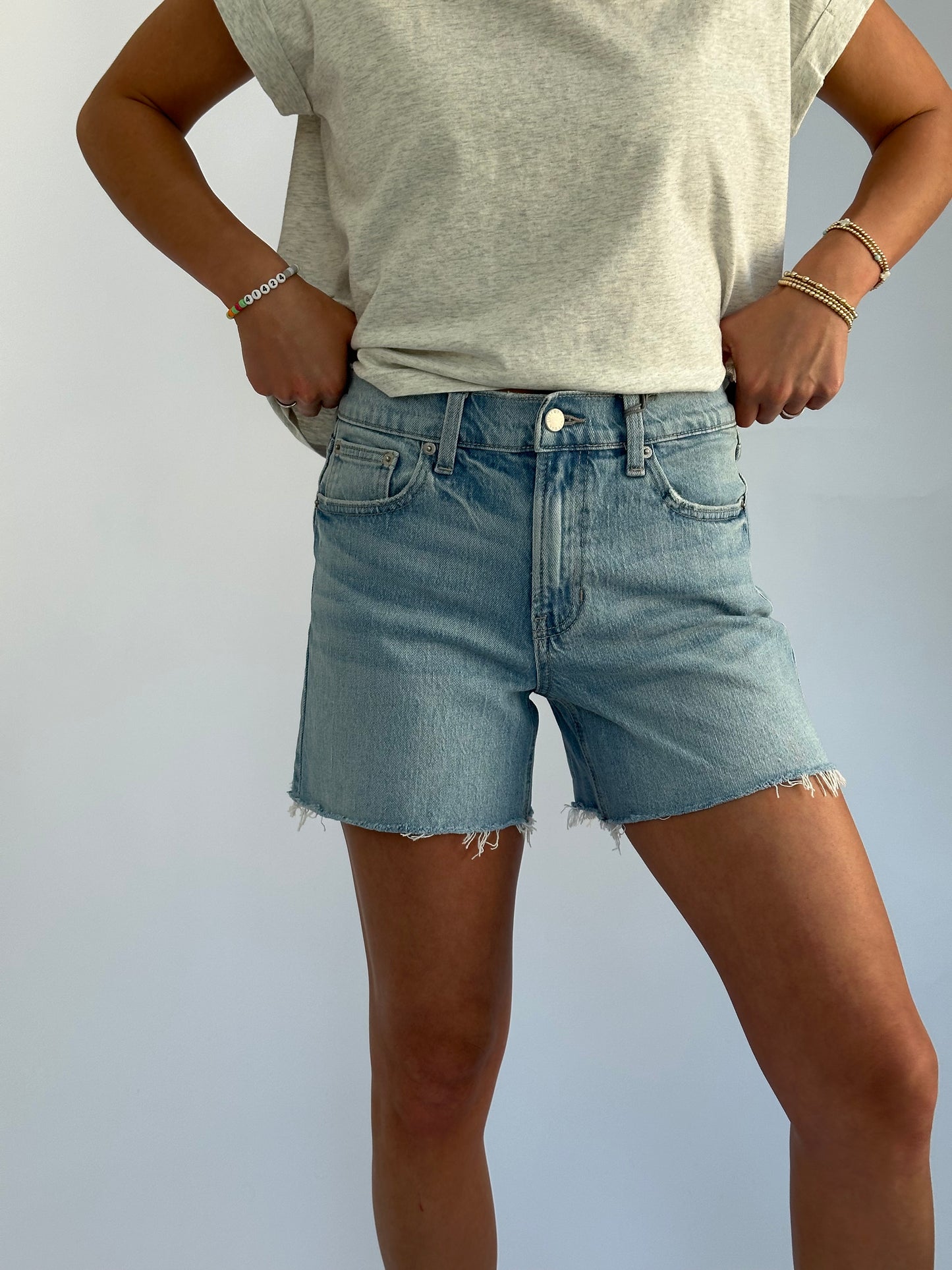 Person wearing a casual gray t-shirt and light blue denim shorts. Hands are on hips, conveying a relaxed and confident vibe against a plain background.