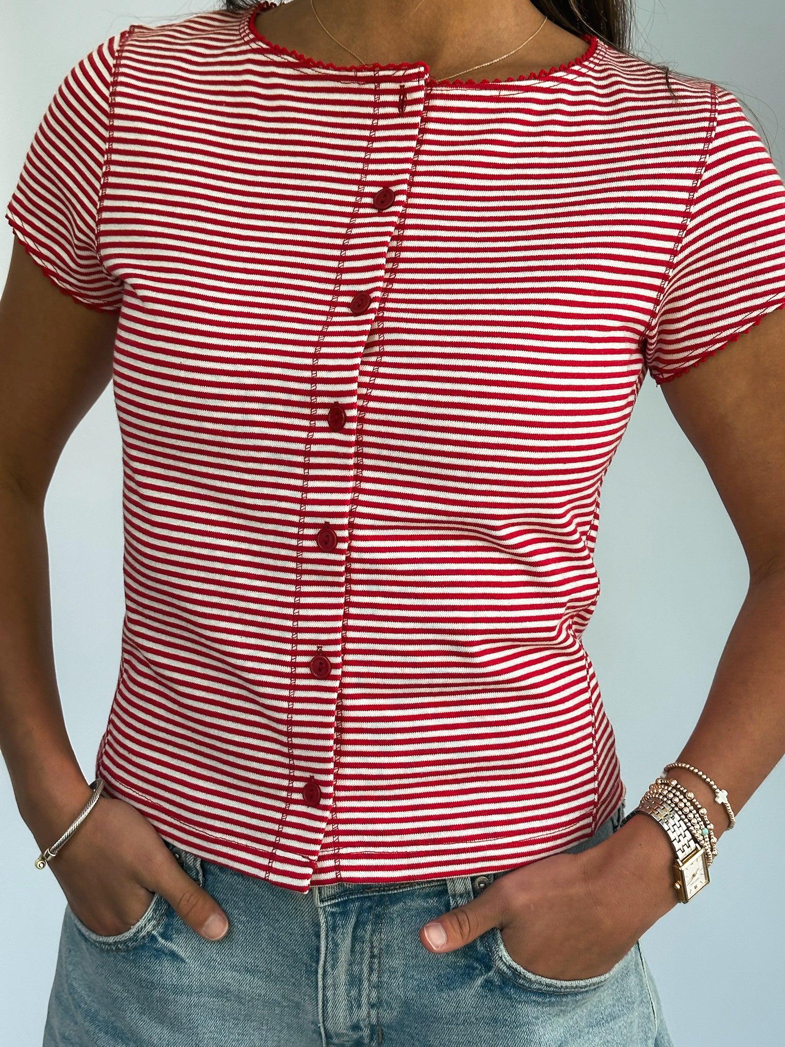 A person wearing a red and white striped button-up shirt with short sleeves and light blue jeans. The hands are in the pockets. The scene is casual.