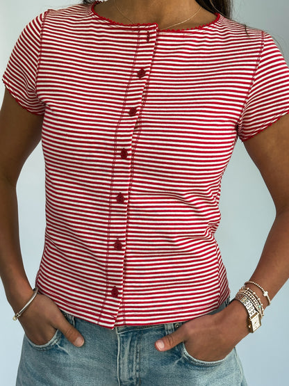 A person wearing a red and white striped button-up shirt with short sleeves and light blue jeans. The hands are in the pockets. The scene is casual.