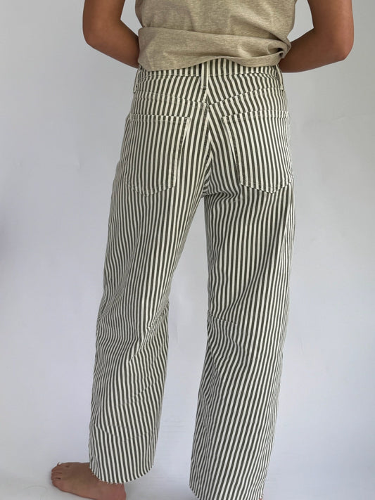 A person wearing cream and olive vertically striped barrel jeans and a beige shirt is holding the shirt hem. The background is plain light gray.