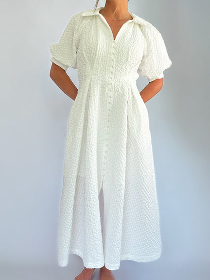 Woman in a textured white dress with buttons stands against a plain backdrop. The dress has short sleeves and a collar, conveying elegance and simplicity.
