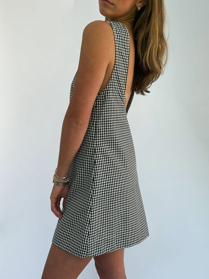 A woman in a sleeveless black and white checkered dress stands sideways against a plain background. Her hair is loose, and she wears a bracelet, exuding a casual yet stylish vibe.