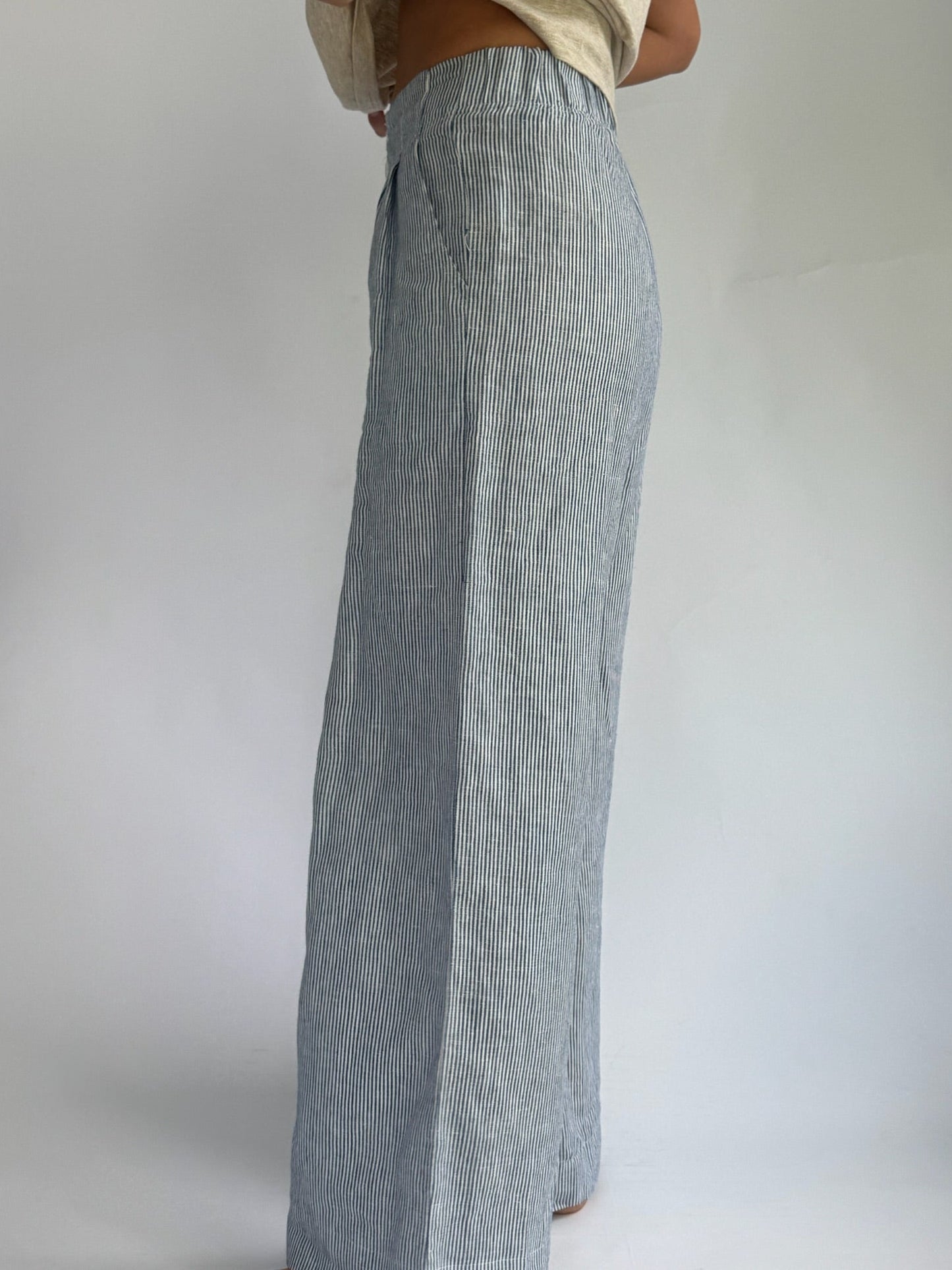 Person wearing high-waisted, blue and white striped, wide-leg linen pants with buttons. Hands on hips; casual, relaxed tone. Barefoot on plain background.