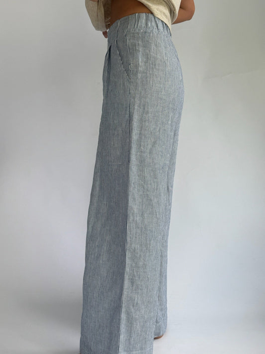 Person wearing high-waisted, blue and white striped, wide-leg linen pants with buttons. Hands on hips; casual, relaxed tone. Barefoot on plain background.