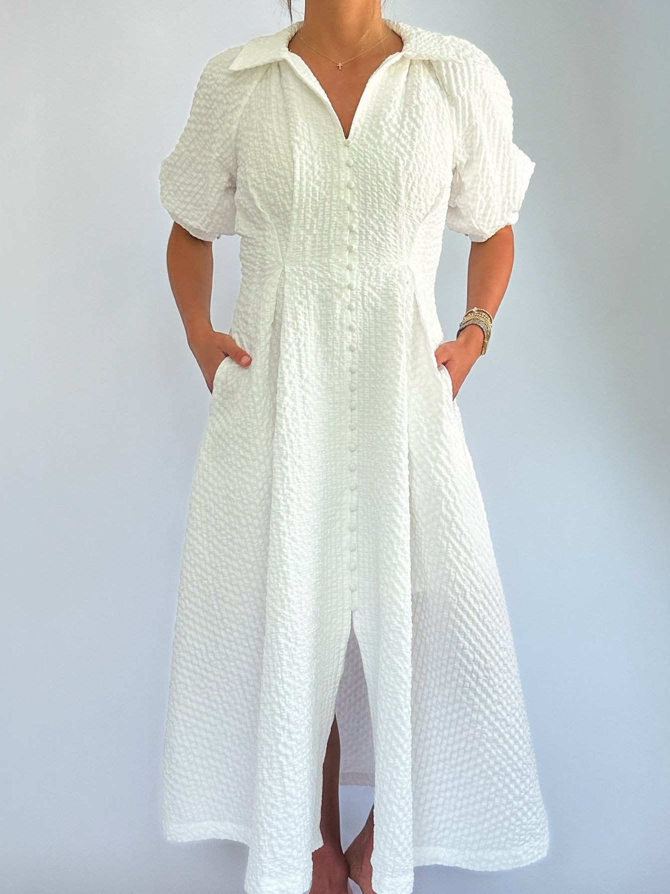 A person wearing a textured white button-up midi dress with short sleeves, hands in pockets; the setting is bright and minimalist.