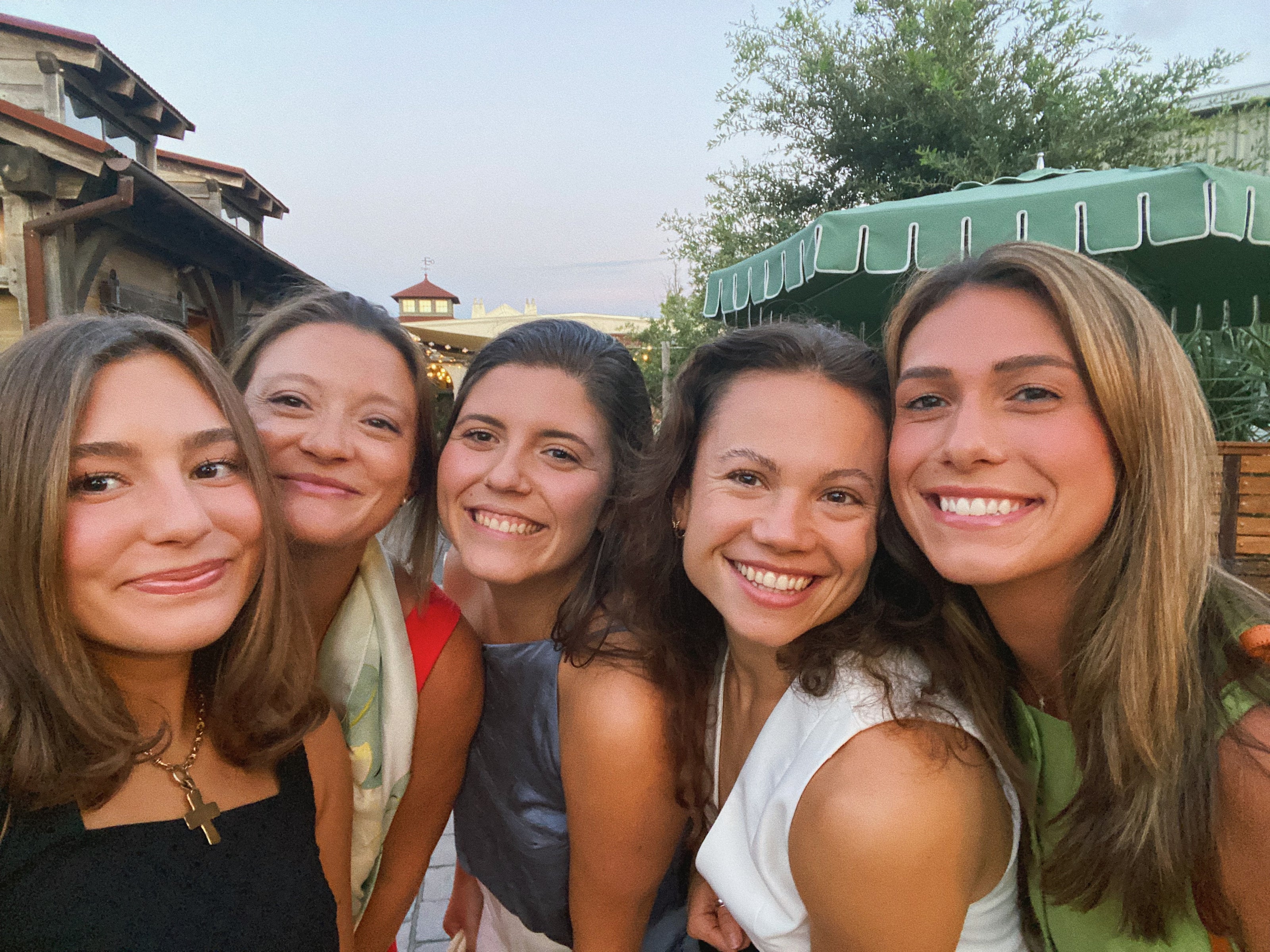 Five young women smiling and posing closely together outdoors. They appear joyful and relaxed, with a backdrop of greenery and a canopy.