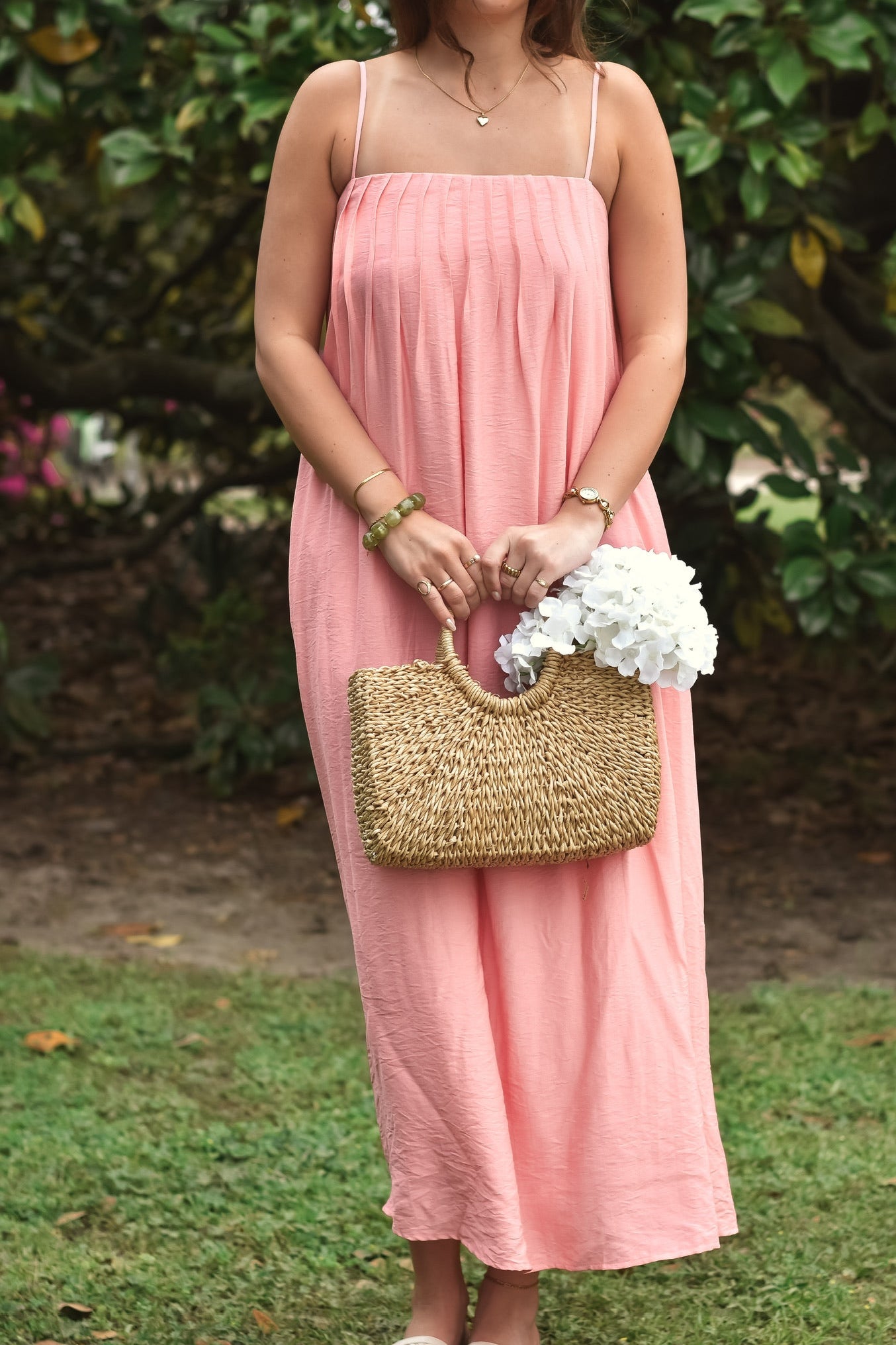 Woman in a pink sundress stands outdoors, holding a woven handbag with white flowers. Lush green foliage in the background adds a serene tone.