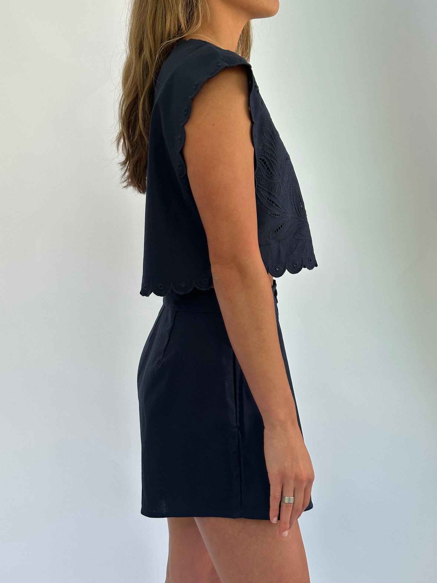 A woman in a navy-blue sleeveless top and skirt is shown from the side. The top has scalloped edges and a textured design. The background is plain white.