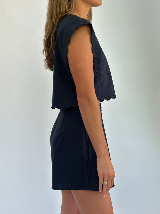 A woman in a navy-blue sleeveless top and skirt is shown from the side. The top has scalloped edges and a textured design. The background is plain white.