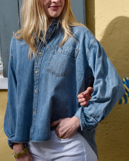 A person wearing a denim shirt and white pants stands against a yellow wall. Their hand, adorned with rings, rests in a pocket, conveying casual elegance.