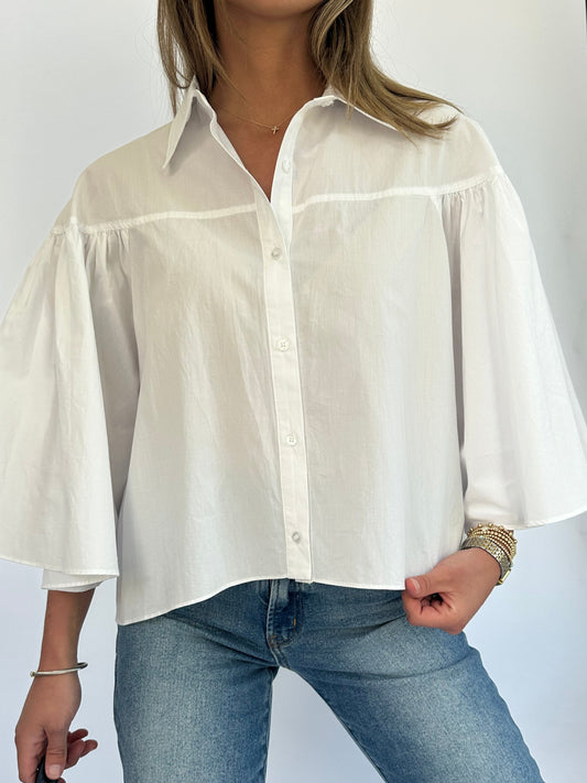 Woman wearing a white button-up shirt with wide, flared sleeves and blue jeans. Her hand holds the bottom of the shirt, displaying gold bracelets.