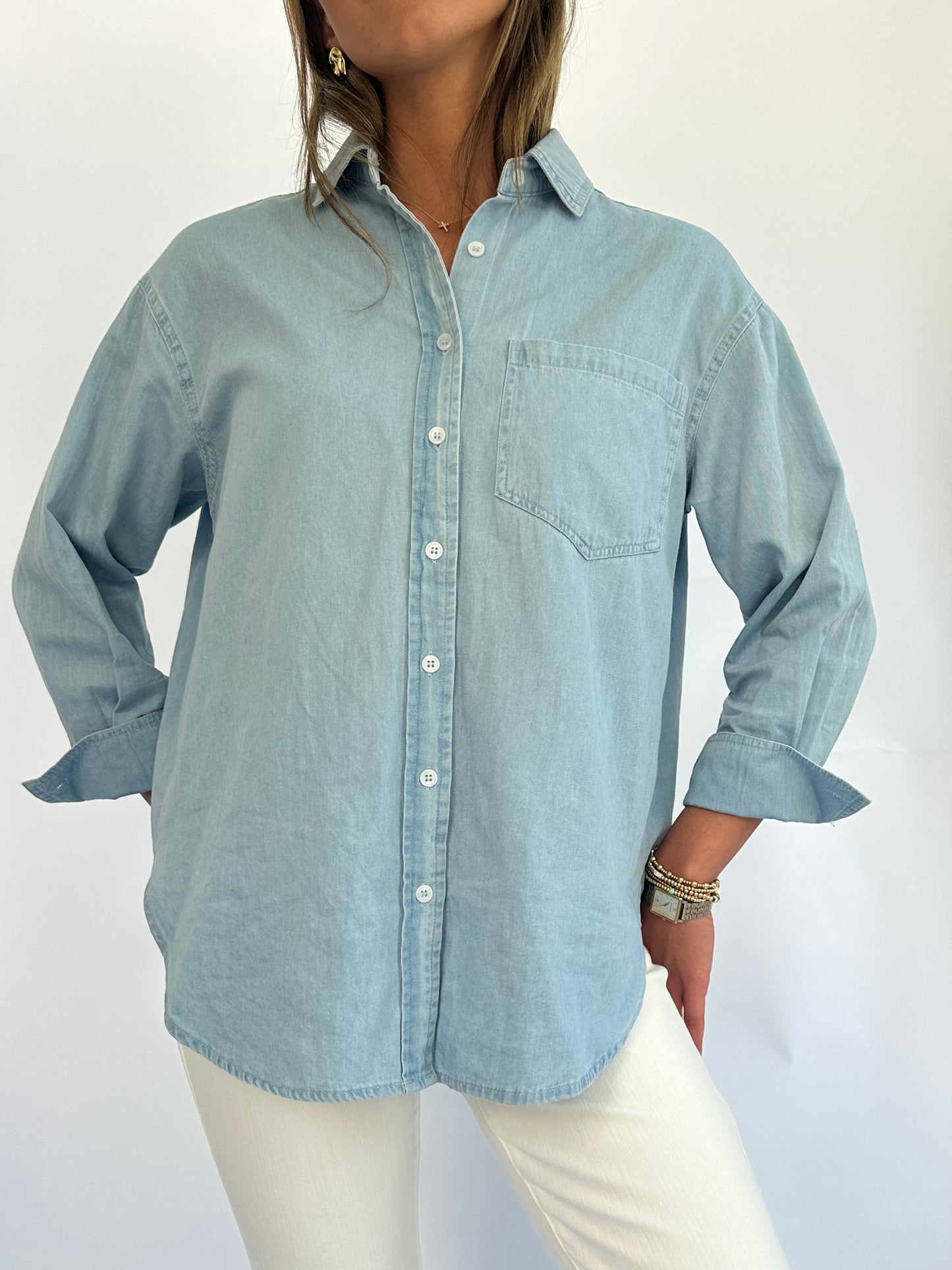 A woman in a light blue denim button-up shirt and white pants is standing against a plain background, exuding a casual, relaxed vibe.