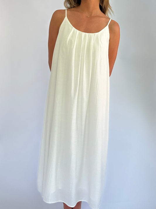 A person wearing a flowing white spaghetti strap dress stands against a plain background. The simple, elegant design conveys a feeling of calm and grace.