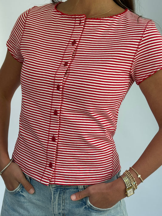 A person wearing a red and white striped button-up shirt and blue jeans, accessorized with gold bracelets and a watch, stands with hands in pockets.