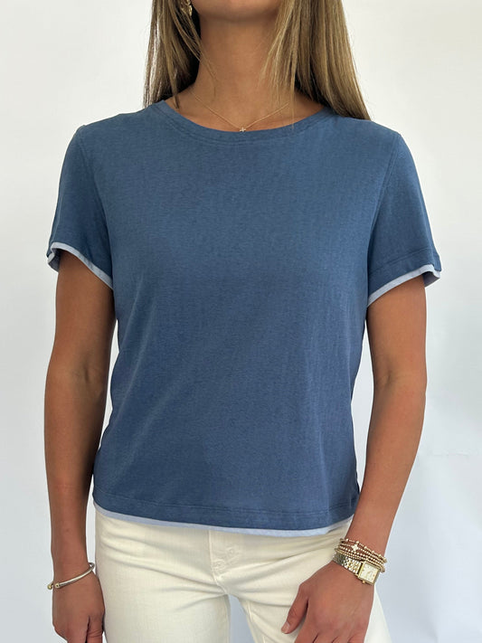 A person wearing a blue short-sleeved T-shirt is shown from the shoulders down against a white background. The outfit is casual and relaxed.